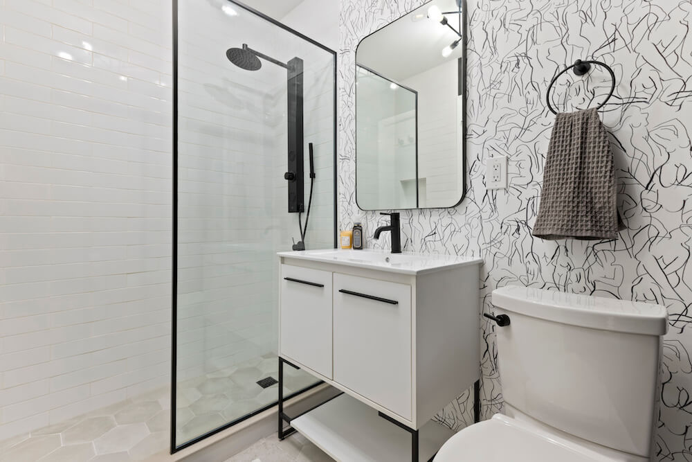 Frameless shower screen with black trim in a modern bathroom featuring bold patterned wallpaper and minimalist fixtures.